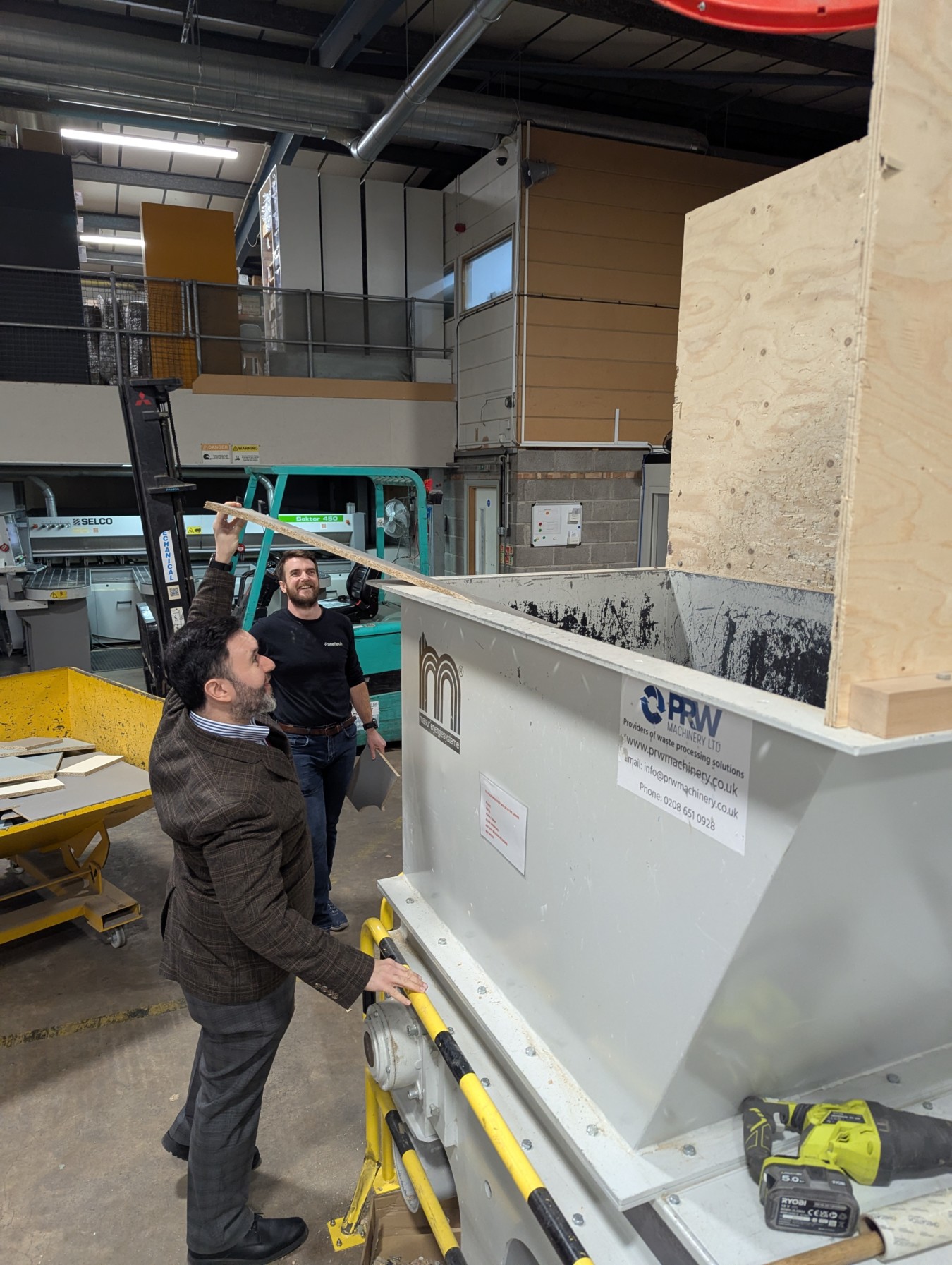Sadik Al-Hassan, Member of Parliament (MP) of the United Kingdom, feeding the wood waste chipper at Paneltech, with Paneltech Director Ross Larner observing in the background. This system processes wood offcuts into biomass fuel for a Ranheat WA 300 industrial heating system.